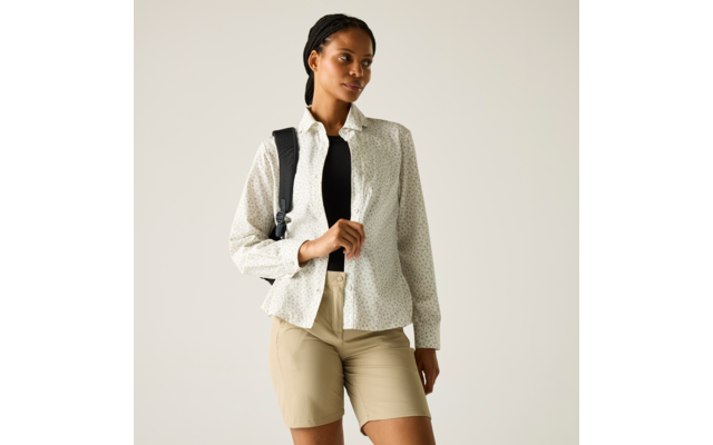 Frau mit geflochtenem Haar, trägt weiße gemusterte Hemdbluse über schwarzem Top, beige Shorts, Rucksackriemen über einer Schulter; steht und blickt zur Seite