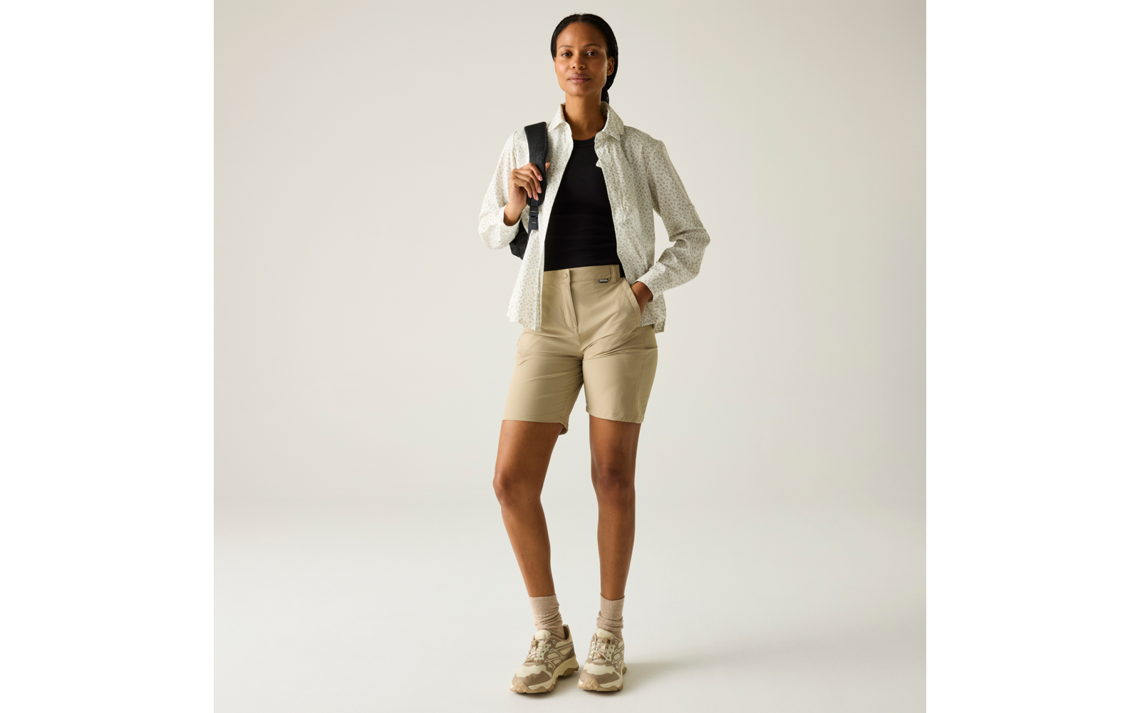 Person steht frontal, trägt helles Hemd über schwarzem Top, beige knielange Shorts, helle Wanderschuhe, Rucksack über einer Schulter, eine Hand in der Tasche