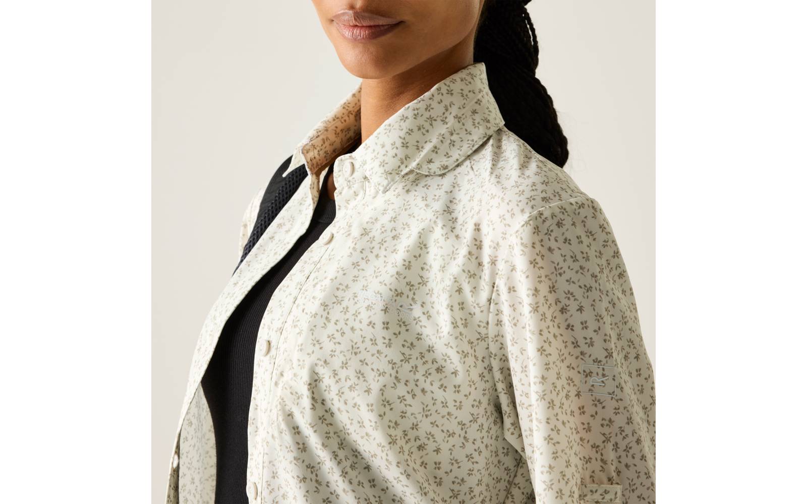 Halbnaher Ausschnitt einer Person von Schultern bis Mund; helles Hemd mit kleinem Blumenmuster und aufgeklapptem Kragen, darunter ein schwarzes Oberteil; geflochtenes Haar hinten sichtbar