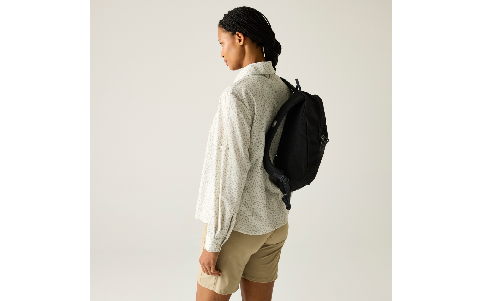 Person im Seitenprofil mit geflochtenem Haar, weißem gemustertem Langarmhemd, beigen Shorts und schwarzem Rucksack vor hellem Hintergrund