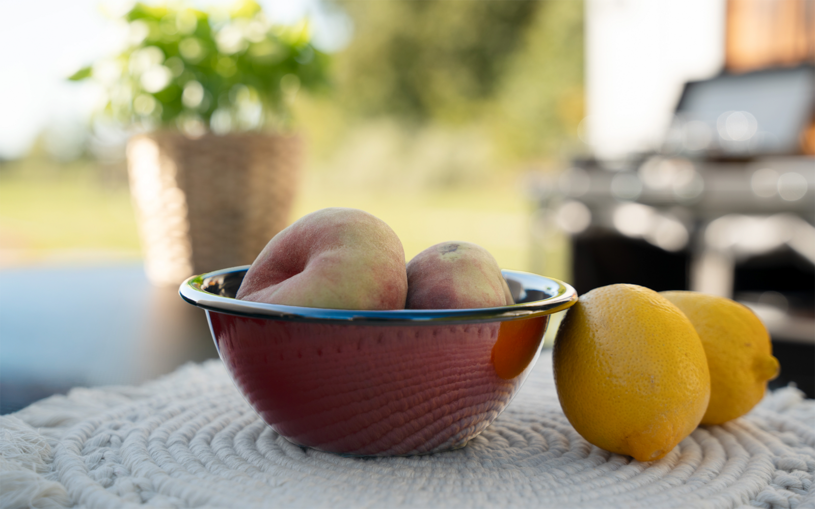 Bol rouge avec des pêches sur une nappe blanche texturée, deux citrons à droite, panier de plante et barbecue flous en arrière‑plan