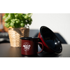 Tasse de camping en émail rouge avec logo blanc à côté d'une tasse retournée, plante de basilic dans un panier tressé en arrière-plan sur une table noire