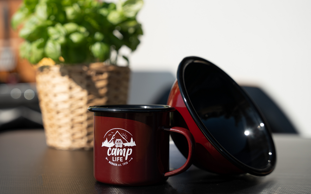Tasse de camping en émail rouge avec logo blanc à côté d'une tasse retournée, plante de basilic dans un panier tressé en arrière-plan sur une table noire