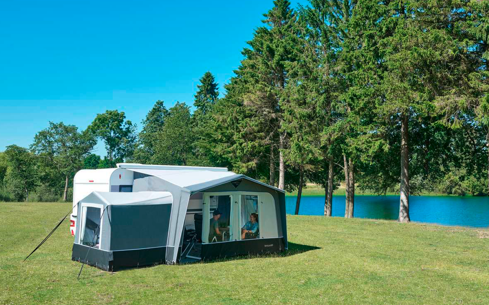 Caravana blanca con toldo gris anexo sobre un campo verde, al fondo pinos altos y un lago azul bajo un cielo despejado