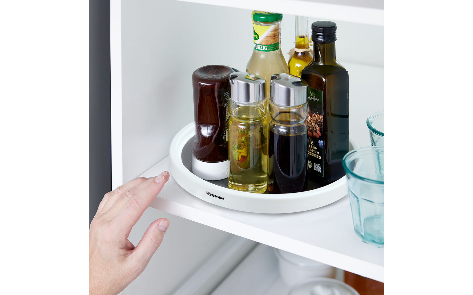 White shelf with a round white turntable holding several glass and plastic bottles with liquids of different colors; two blue drinking glasses on the right; a hand on the left touching the turntable