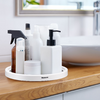 Round white tray labeled Westmark on a wooden countertop; on it several bottles and jars: white pump bottle with black pump, brown spray bottle with black trigger, white cream jar, several white bottles; in the background a white sink and a mirror