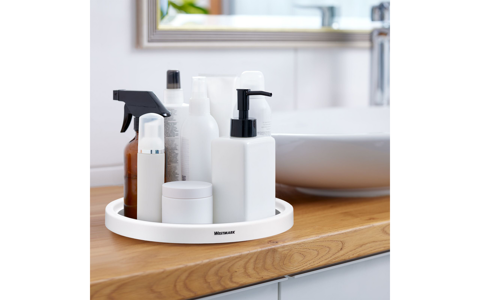 Round white tray labeled Westmark on a wooden countertop; on it several bottles and jars: white pump bottle with black pump, brown spray bottle with black trigger, white cream jar, several white bottles; in the background a white sink and a mirror