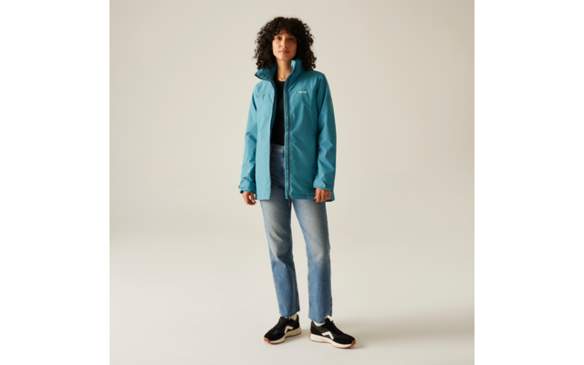 Femme aux cheveux bouclés portant une veste turquoise, un jean bleu et des chaussures noires, debout devant un fond clair