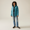 Femme aux cheveux bouclés portant une veste turquoise, un jean bleu et des chaussures noires, debout devant un fond clair