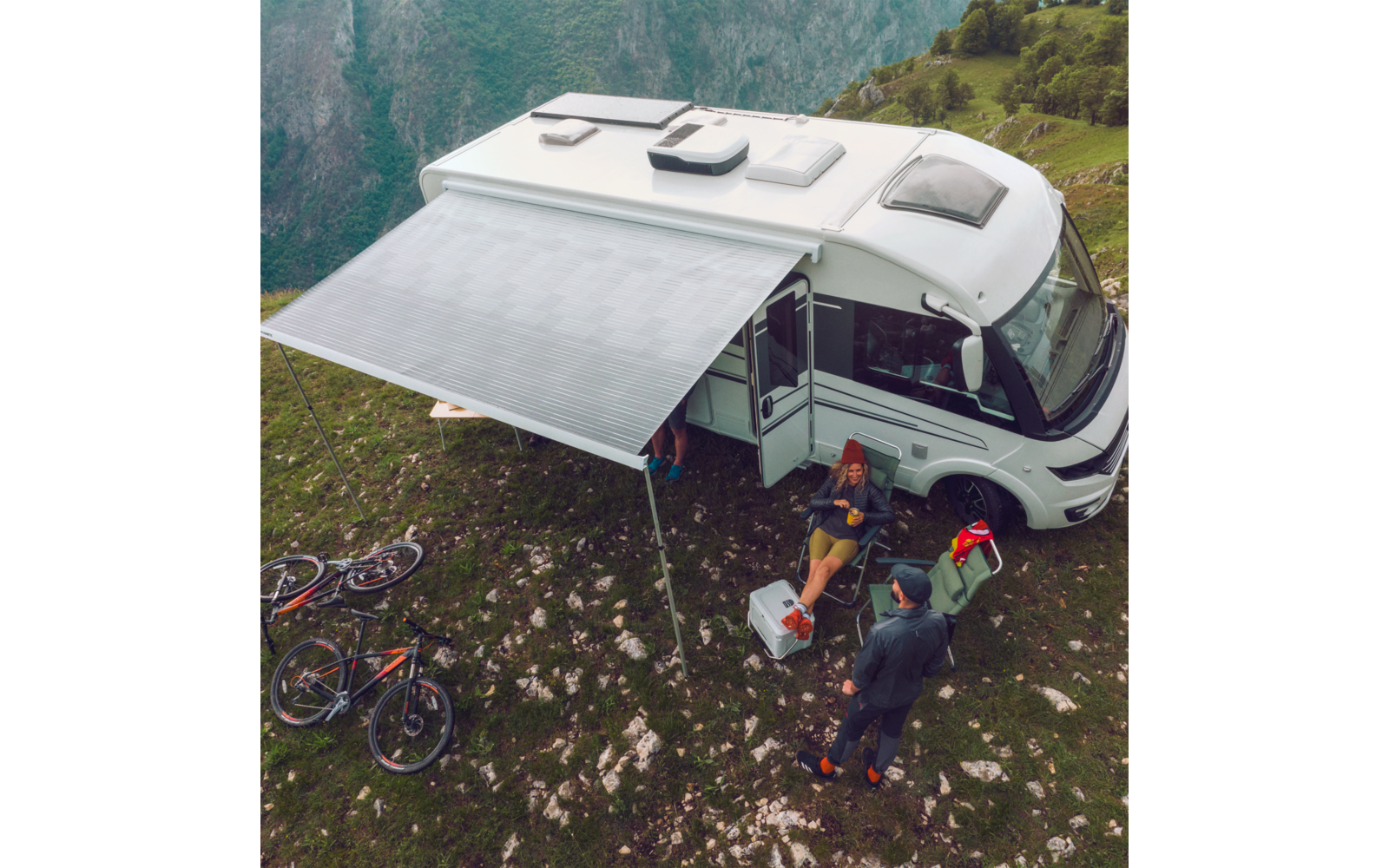 Camper parcheggiato su terreno erboso con tenda da sole aperta, due biciclette appoggiate al veicolo, persona seduta davanti al camper