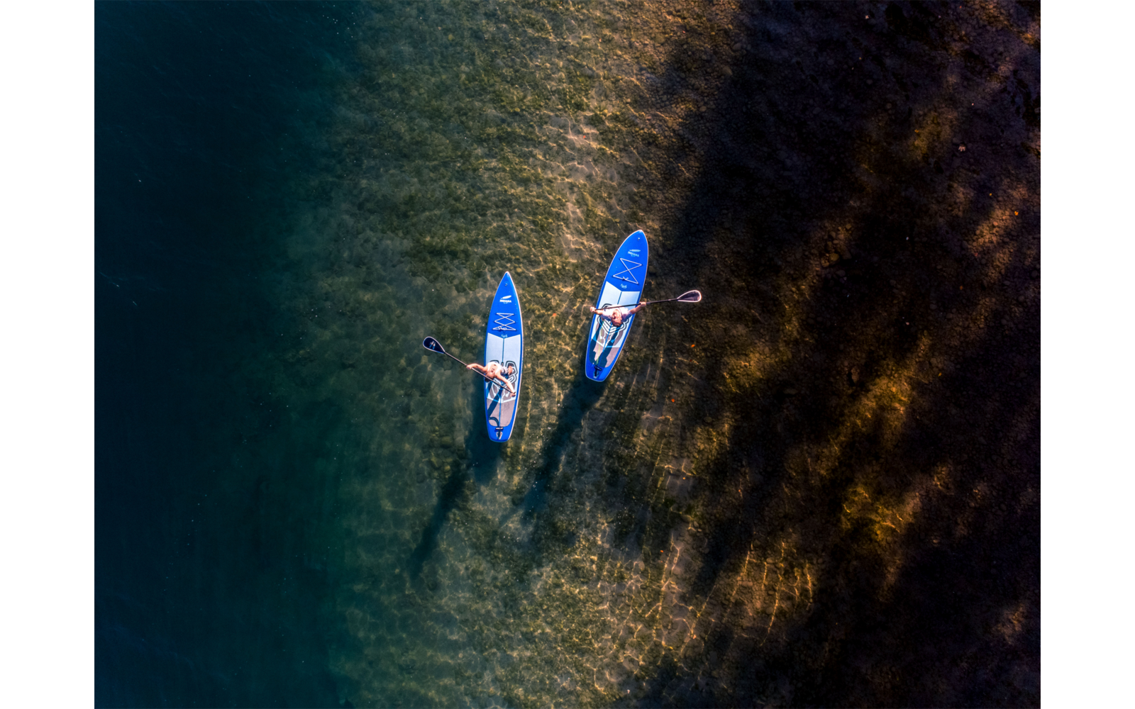 Luftaufnahme von zwei Personen auf blauen Stand-up-Paddleboards mit Paddeln über flachem Wasser, neben dunklen Algenflächen und sonnenbeleuchteten Wasserreflexen