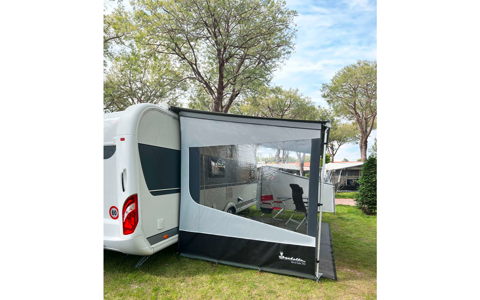 Caravana blanca con toldo anexo con gran ventana transparente; dentro del toldo dos sillas de camping y una mesa pequeña; árboles y césped en el fondo