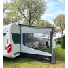 Caravana blanca con toldo anexo con gran ventana transparente; dentro del toldo dos sillas de camping y una mesa pequeña; árboles y césped en el fondo