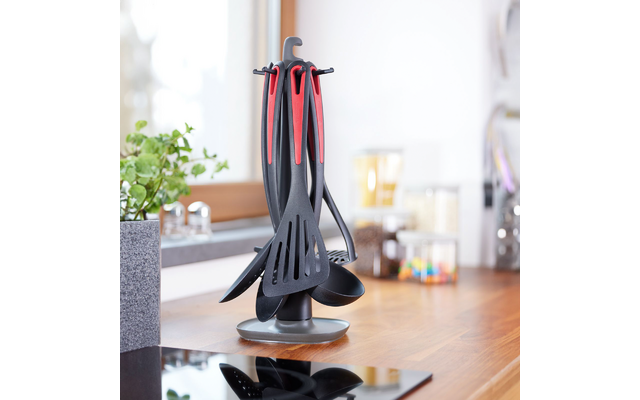 Utensilios de cocina negros con mangos rojos en un soporte giratorio sobre encimera de madera; fondo de cocina desenfocado