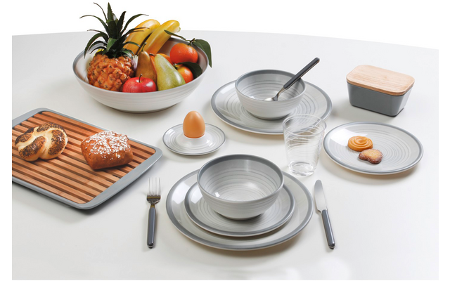 Weißer Tisch mit grauen Tellern und Schüsseln, Löffel in einer Schüssel, Glas, Messer und Gabel, Eierbecher mit Ei, große Obstschale mit Ananas, Bananen, Äpfeln und Birnen, Holzbrett mit Brötchen und Laugengebäck, kleine Platte mit Keksen, rechteckig