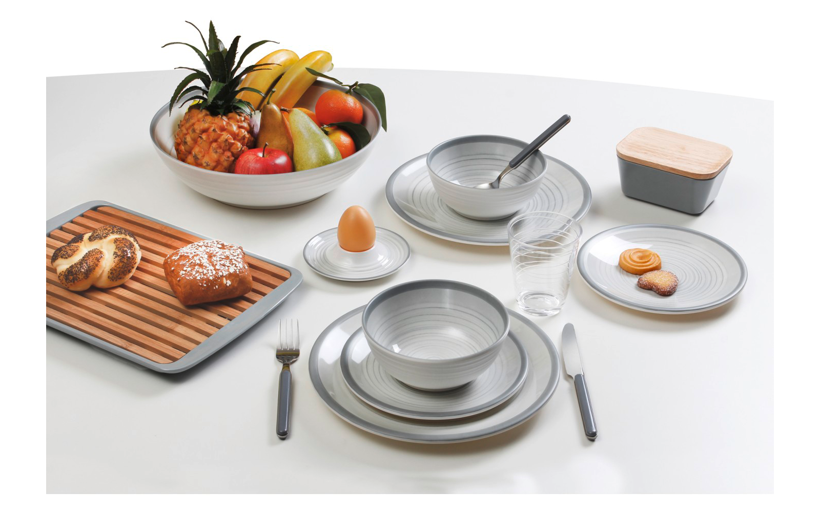 Weißer Tisch mit grauen Tellern und Schüsseln, Löffel in einer Schüssel, Glas, Messer und Gabel, Eierbecher mit Ei, große Obstschale mit Ananas, Bananen, Äpfeln und Birnen, Holzbrett mit Brötchen und Laugengebäck, kleine Platte mit Keksen, rechteckig
