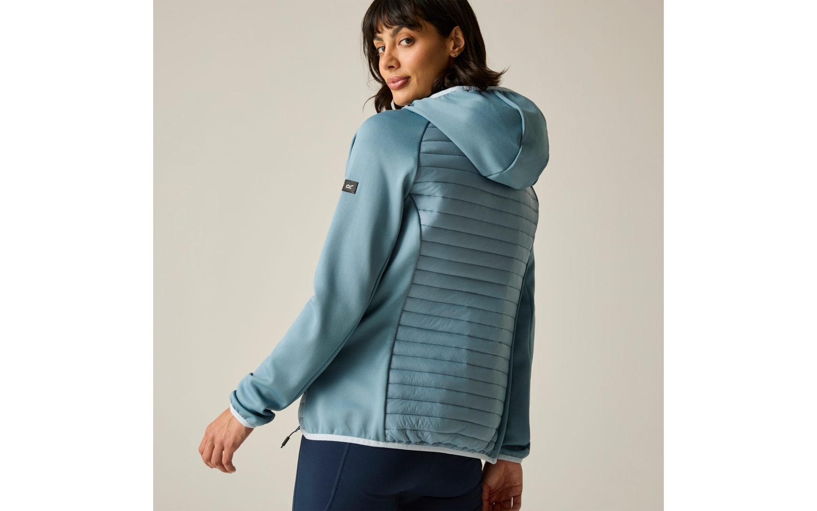 Woman in three-quarter back view looking over her shoulder, wearing a light blue hooded jacket with a quilted back panel and dark pants, neutral background