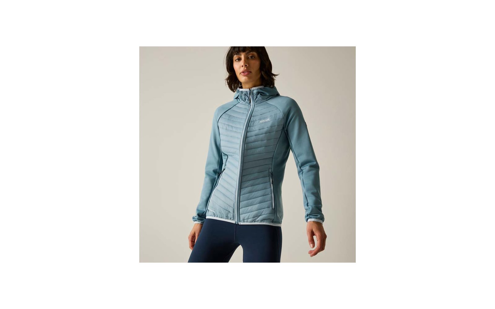 Person wearing a light blue quilted hooded jacket with full zipper and zippered pockets, dark leggings, plain background