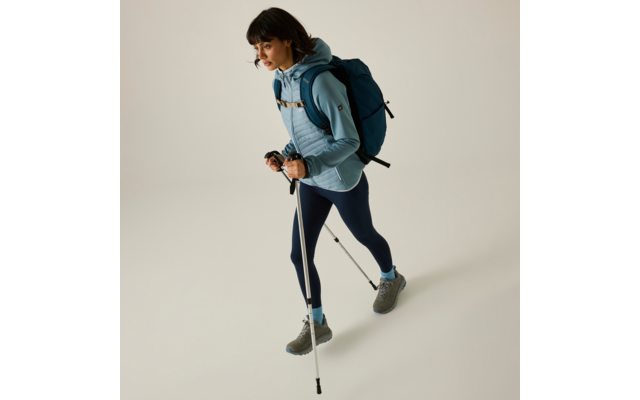 Side view of a person in outdoor clothing: light blue hooded jacket, dark leggings, gray shoes, blue backpack, holding two trekking poles; plain light background