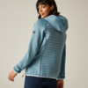 Woman in three-quarter back view looking over her shoulder, wearing a light blue hooded jacket with a quilted back panel and dark pants, neutral background
