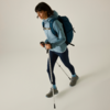 Side view of a person in outdoor clothing: light blue hooded jacket, dark leggings, gray shoes, blue backpack, holding two trekking poles; plain light background