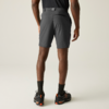 Person von hinten, knielange dunkelgraue Shorts, schwarzes T‑Shirt, schwarze Socken und schwarze Schuhe mit orangefarbenen Details, Hände seitlich, heller einfarbiger Hintergrund