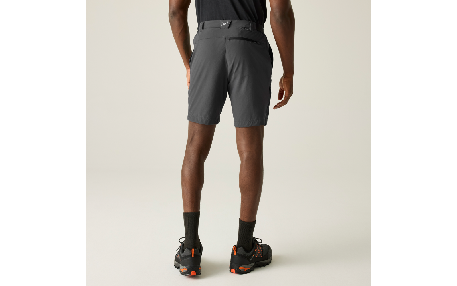 Person von hinten, knielange dunkelgraue Shorts, schwarzes T‑Shirt, schwarze Socken und schwarze Schuhe mit orangefarbenen Details, Hände seitlich, heller einfarbiger Hintergrund