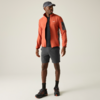 Stehende Person in orangefarbener Jacke, schwarzem T‑Shirt, grauen Shorts, grauem Cap, schwarzen Socken und schwarzen Schuhen mit orangefarbenen Details vor hellem Hintergrund