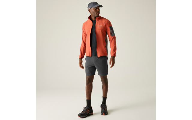 Stehende Person in orangefarbener Jacke, schwarzem T‑Shirt, grauen Shorts, grauem Cap, schwarzen Socken und schwarzen Schuhen mit orangefarbenen Details vor hellem Hintergrund