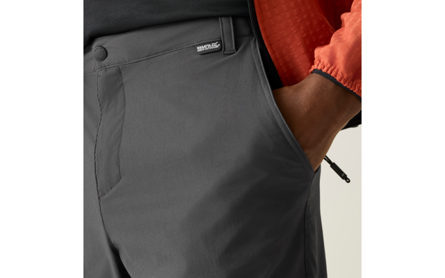 Nahaufnahme des Hüftbereichs einer Person mit grauer Hose und orangefarbener Jacke, rechte Hand in Hosentasche, kleines Markenlabel am Hosenbund sichtbar