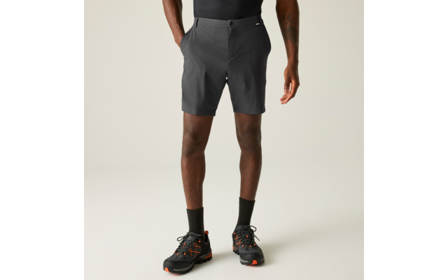 Körper eines Mannes von der Taille abwärts, dunkle Haut; er trägt dunkelgraue Shorts, schwarze Socken und schwarze Sportschuhe mit orangefarbenen Akzenten; Hände in den Taschen; heller einfarbiger Hintergrund