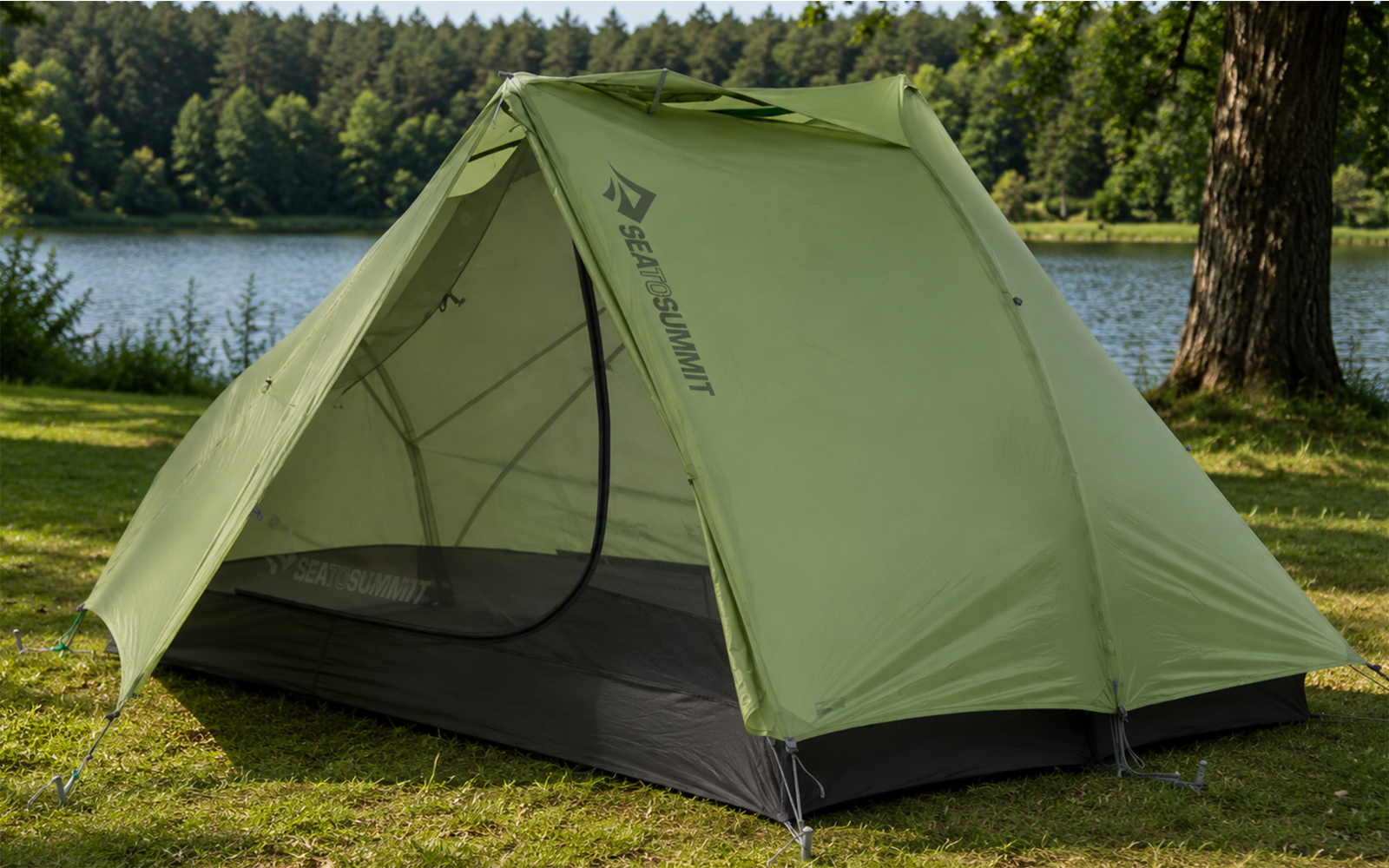 Green tent with open front flap and visible mesh inner door, pitched on grass, lake and line of trees in the background, tree trunk visible at right