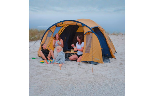Vier Personen sitzen vor einem gelb-blauen Zelt auf einem Sandstrand; Spielzeug im Vordergrund; Meer und Dünen im Hintergrund