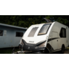 Caravana blanca con logo KNAUS negro, ventana frontal, enganche de remolque y ventana lateral; al fondo otra caravana y árboles