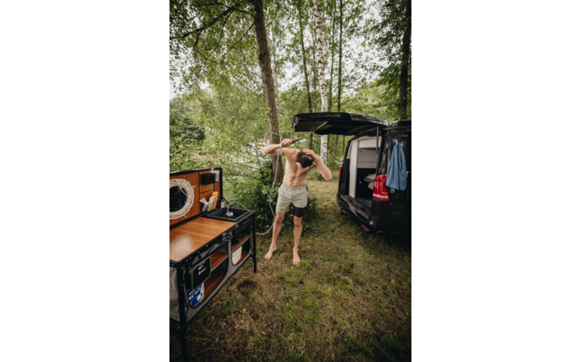 Person ohne Oberteil, barfuß, trägt helle Shorts und hält ein Tuch über dem Kopf, steht neben offenem schwarzem Camper-Van mit Klappküche; Bäume und Wasser im Hintergrund