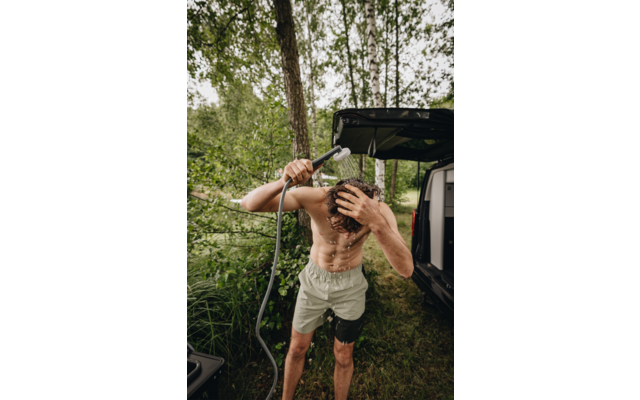 Person ohne Oberteil hält Gartenschlauch und gießt Wasser über den Kopf neben offenem Camper im Wald, trägt kurze Hose und Kniebandage