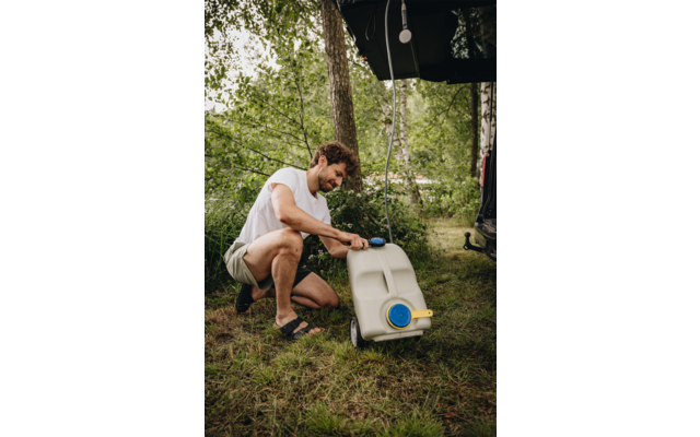Mann hockt neben weißem Wassertank mit blauem Verschluss und gelbem Ausguss; im Hintergrund Bäume, Gras und ein offenes Fahrzeugheck mit hängender Lampe