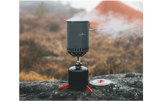 Grauschwarzer, zylindrischer Kochtopf auf schwarzem Gasbehälter mit roten Standfüßen, Dampf steigt auf; runder Deckel liegt daneben auf einem Felsen; unscharfer Hintergrund mit orangefarbenem Zelt