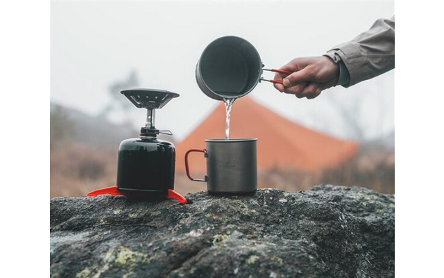 Hand gießt Wasser aus einem Metallbecher in eine Metalltasse auf einem Felsen; daneben ein Gaskocher; im Hintergrund ein orangefarbenes Zelt