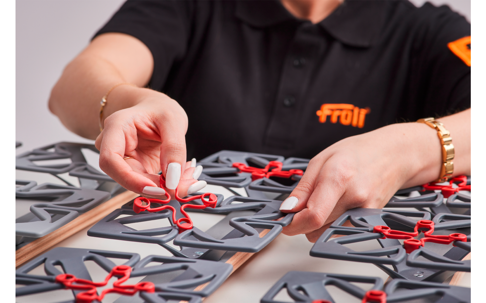 Hände über grauen Kunststoffplatten mit roten Verbindungsteilen auf einem Tisch; schwarzes Poloshirt mit orangefarbenem Logo sichtbar