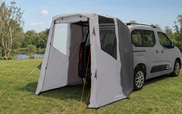 Light gray van with open sliding door; an attached light-gray awning/tent with windows and orange guy lines; grass, lake, trees and blue sky with clouds in the background