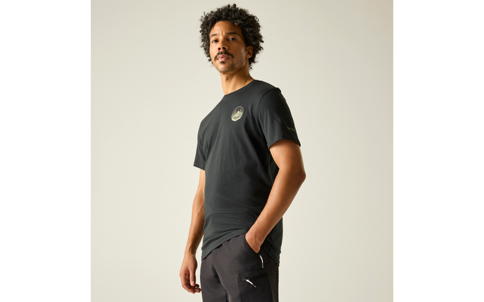 Mann steht leicht seitlich, trägt schwarzes T‑Shirt mit rundem Brustlogo und schwarze Hose, eine Hand in Hosentasche, heller einfarbiger Hintergrund