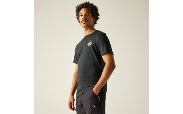 Mann steht leicht seitlich, trägt schwarzes T‑Shirt mit rundem Brustlogo und schwarze Hose, eine Hand in Hosentasche, heller einfarbiger Hintergrund