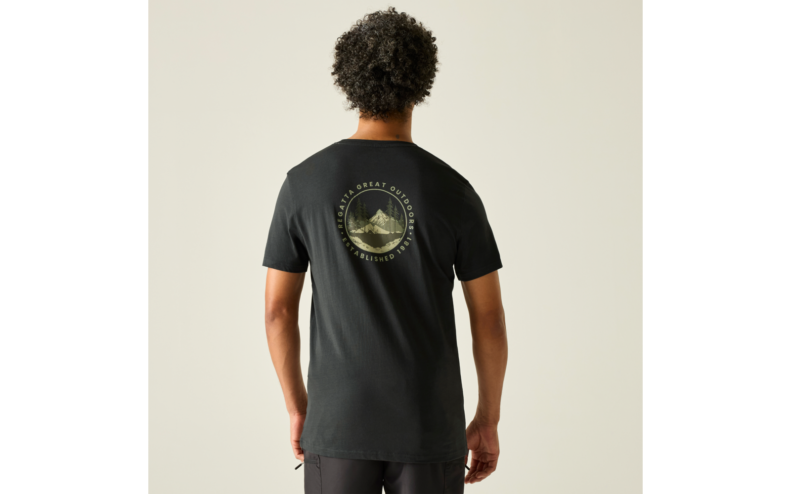 Person von hinten mit dunkel lockigem Haar, trägt schwarzes T‑Shirt mit rundem Aufdruck einer Berglandschaft auf dem oberen Rücken, heller einfarbiger Hintergrund