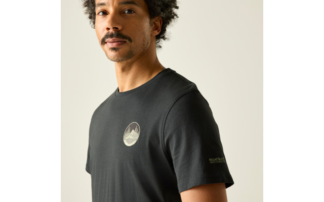 Oberkörper eines Mannes mit lockigem Haar und Schnurrbart, trägt ein schwarzes T‑Shirt mit rundem Berglogo auf der Brust vor hellem Hintergrund