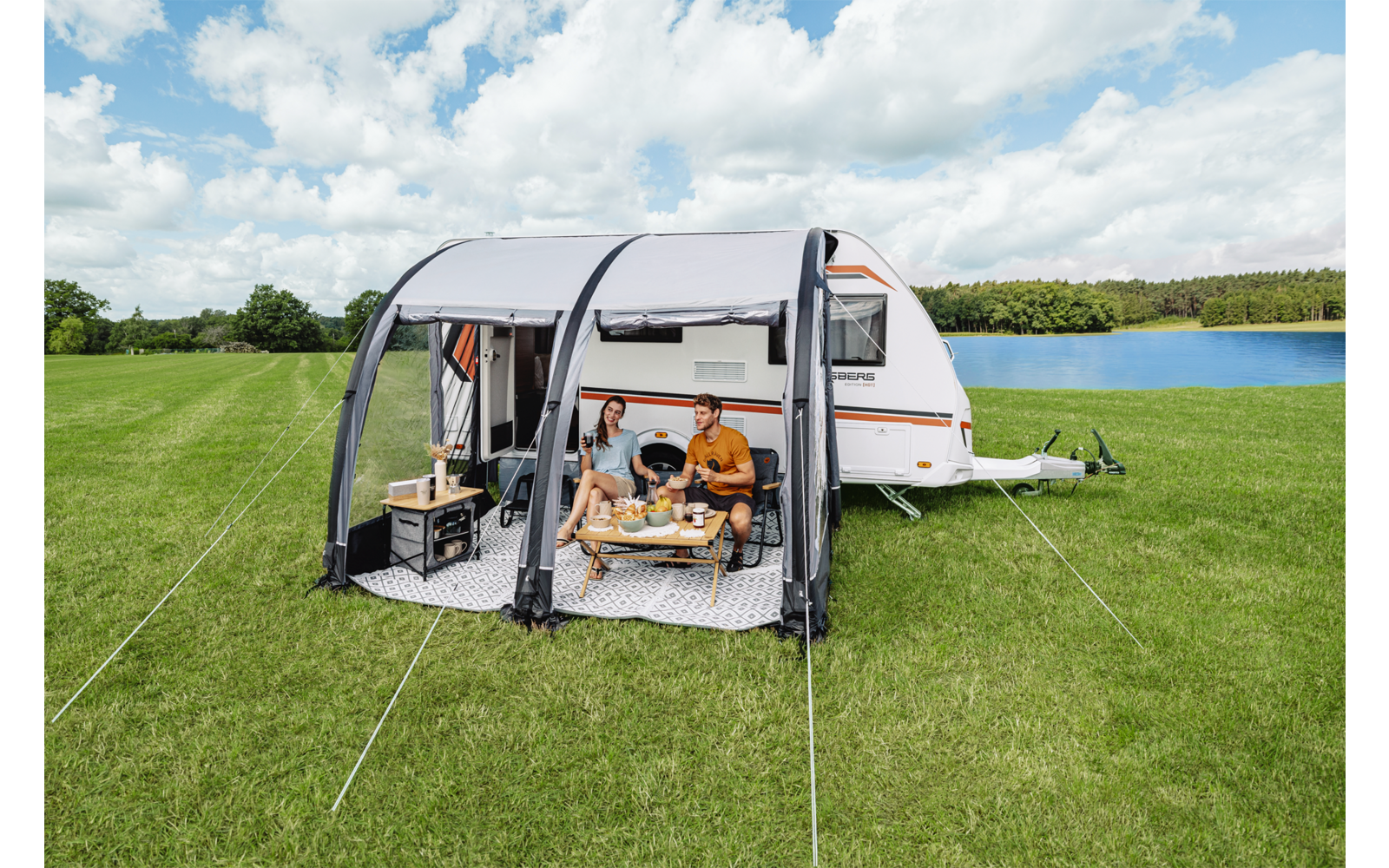 Wohnwagen mit angebautem Vorzelt auf einer Wiese neben einem See; zwei Personen sitzen an einem niedrigen Tisch im Vorzelt; Tisch mit Essen/Getränken, Campingutensilien und Kisten davor; Bäume und wolkiger Himmel im Hintergrund
