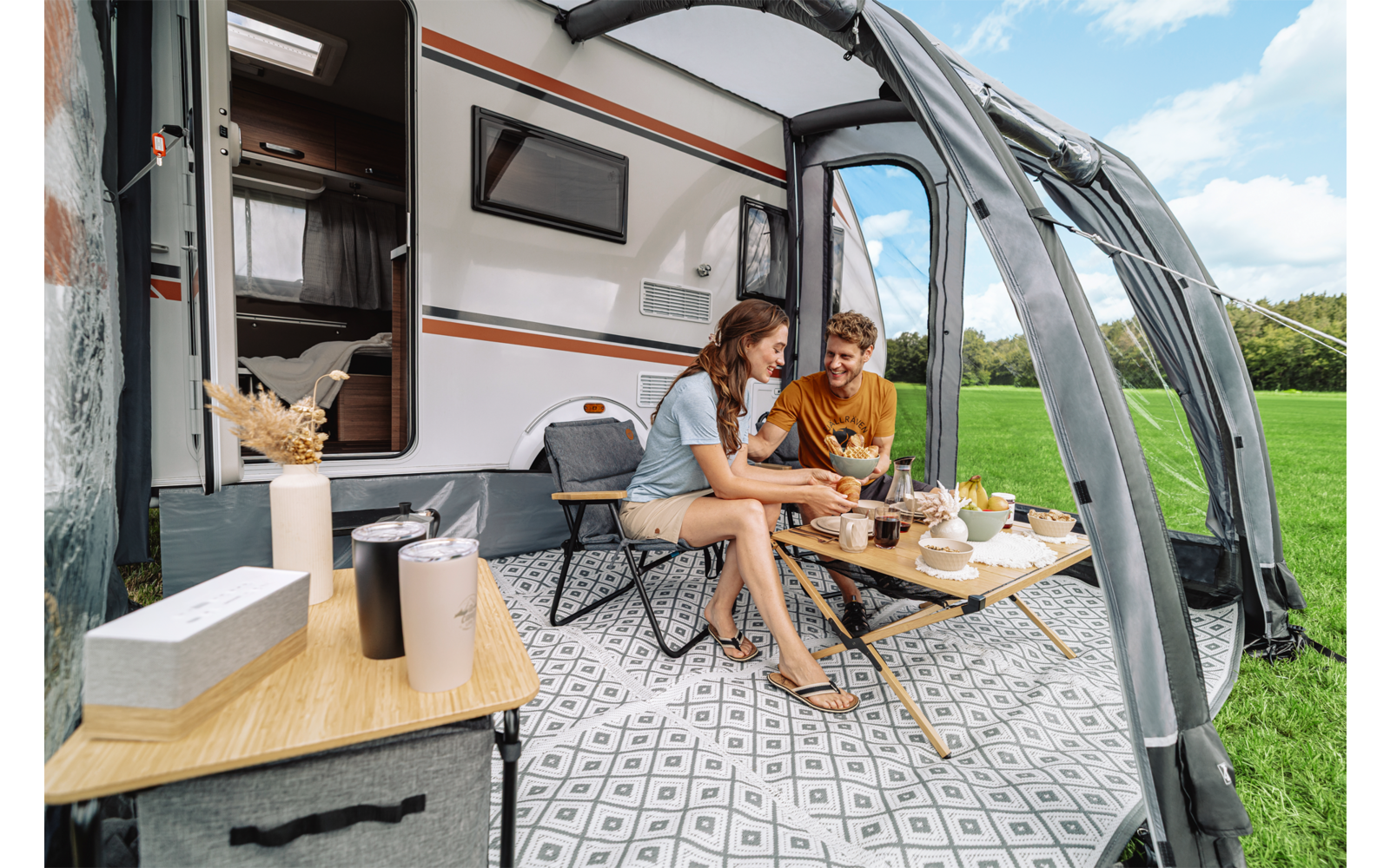 Zwei Personen sitzen an einem niedrigen Tisch im Vorzelt eines Wohnmobils; auf dem Tisch Tassen, Teller, eine Kanne und Snacks; offene Wohnmobiltür, grünes Feld, blauer Himmel und gemusterter Bodenbelag sichtbar