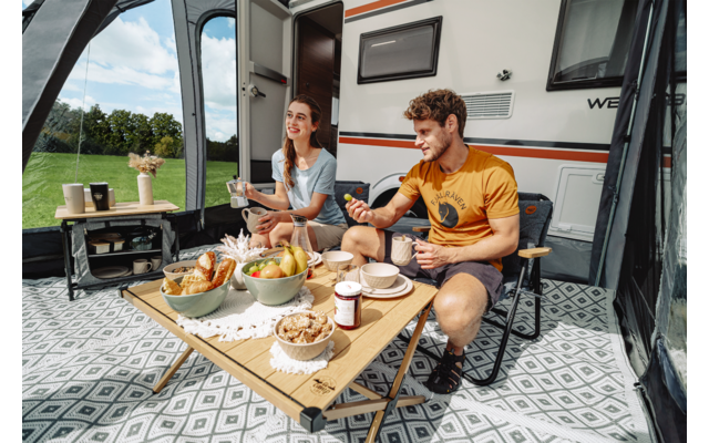 Ein Mann und eine Frau sitzen im Vorzelt eines Wohnmobils an einem niedrigen Tisch mit Obstschale, Brot, Marmelade und Getränken; Wiese im Hintergrund