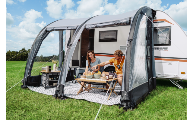 Zwei Personen sitzen an einem niedrigen Tisch im aufgestellten Vorzelt neben einem Wohnwagen auf Gras, blauer Himmel mit Wolken