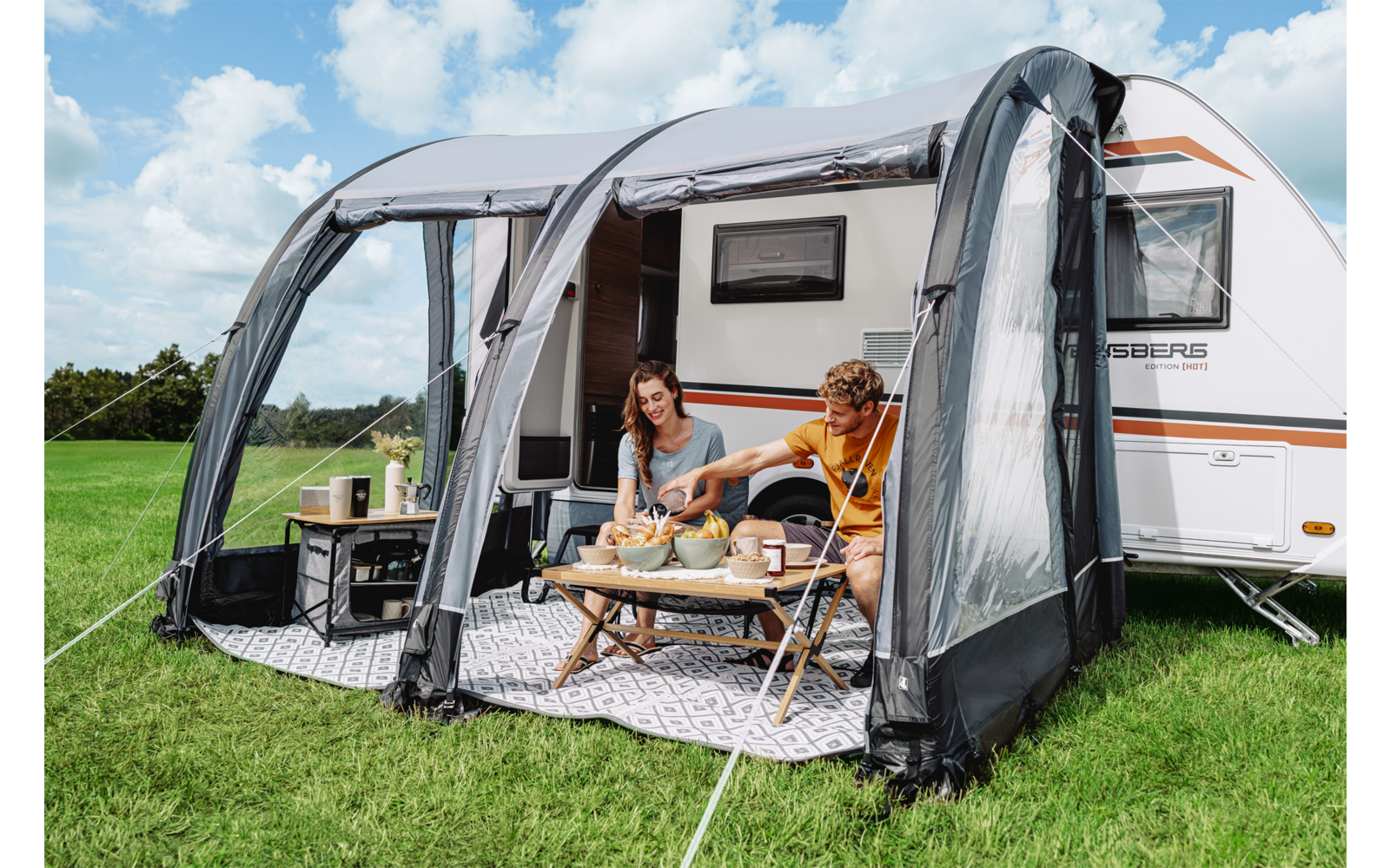 Zwei Personen sitzen an einem niedrigen Tisch im aufgestellten Vorzelt neben einem Wohnwagen auf Gras, blauer Himmel mit Wolken
